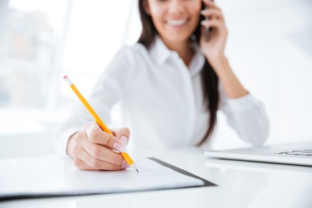 Cropped image of Business woman talking at phone and writing something in documents by the table in office. Focus on documentsの写真素材