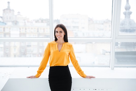 Business woman in orange shirt standing near the window in office and looking at cameraの写真素材