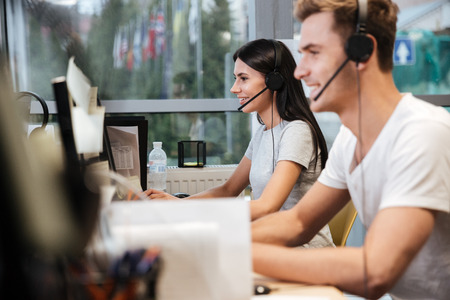 Side view of Coworkers in white t-shirts sit by the table in headphones. Call center.