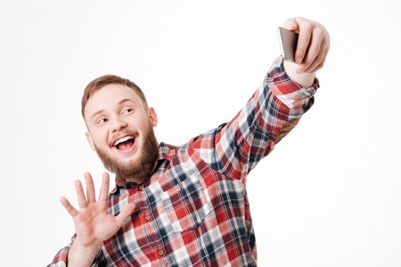 Bearded man in shirt making selfie and waving at smartphone in studio. Isolated white backgroundの写真素材