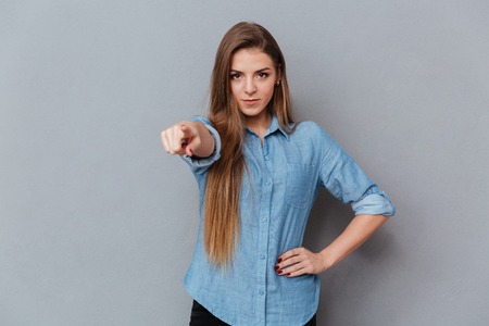 Woman in shirt pointing at camera with arm at hip. Isolated gray background
