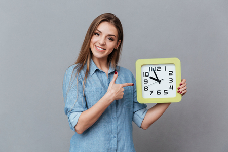 Smiling Woman in shirt holding clock in hand and pointing on them. Isolated gray backgroundの写真素材