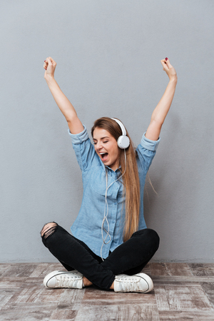 Vertical image of Woman in shirt listening to music on the wooden floor with hands up. Isolated gray backgroundの写真素材