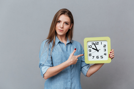 Serious Woman in shirt holding clock in hand and pointing on them. Isolated gray backgroundの写真素材