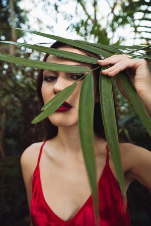 Vertical image of brunette woman in red dress which hiding behind the plant in greenhouse. Close up portraitの写真素材
