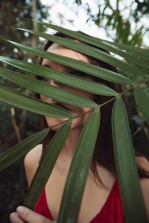 Vertical image of woman in red dress which hiding behind the plant in greenhouse. Close up portraitの写真素材