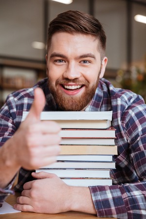 Smiling male student holding stack of books while sitting in library and showing thumbs up gestureの写真素材