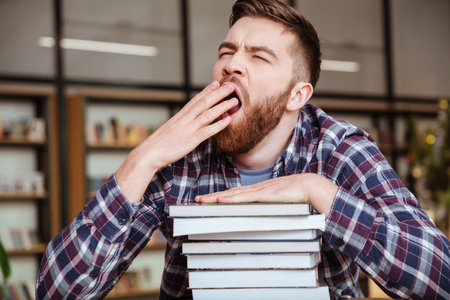 Portrait of a young tired male student yawning while sitting at the library desk with stack of booksの写真素材