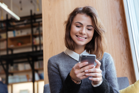 Happy young female student sitting at table and reading text message on her cell phoneの写真素材