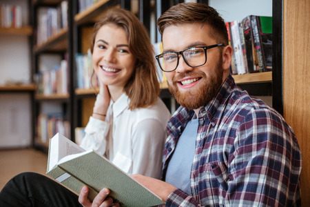 Happy young couple smiling and reading book in libraryの写真素材