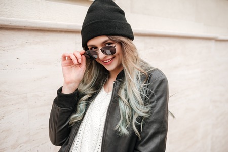 Image of young cheerful lady wearing hat and sunglasses walking on the street.の写真素材