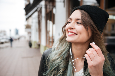 Image of beautiful young woman wearing hat walking on the street and looking aside while listening music with earphones.の写真素材