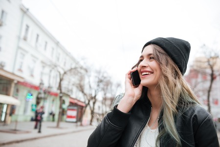 Photo of smiling young woman wearing hat walking on the street and talking by mobile phone.の写真素材