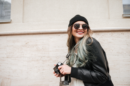 Photo of young smiling lady wearing hat and sunglasses walking on the street while holding camera.の写真素材