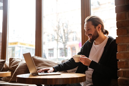 Happy Bearded man sitting by the table with cup of coffee and using laptop in cafeの写真素材