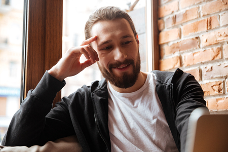 Close up portrait of Smiling Bearded man sitting by the table and using laptop in cafeの写真素材