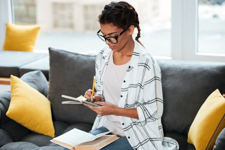 Image of young attractive african lady wearing glasses sitting in library holding book and writing...
