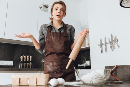 Image of young cheerful woman standing in kitchen and cooking the dough. Looking at camera.