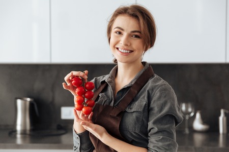 Picture of young happy woman standing in kitchen holding tomatoes in hands.の写真素材