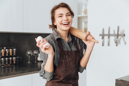 Photo of young laughing lady standing in kitchen and cooking the dough. Looking at camera.の写真素材