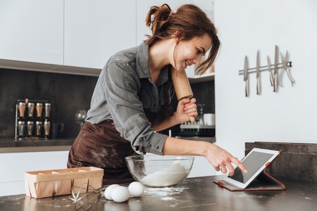 Image of young pretty lady standing in kitchen and cooking the dough. Looking at tablet computer.の写真素材