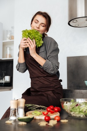 Photo of young woman standing in kitchen smells lettuce leaves and cooking with the tomatoes and avocado.の写真素材