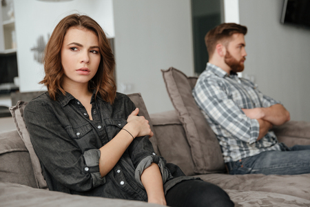 Picture of young sad quarrel loving couple sitting on sofa indoors. Looking aside.の写真素材