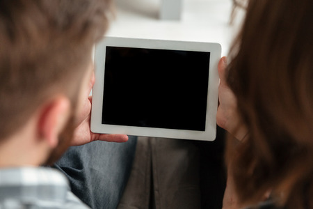 Back view photo of young loving couple in kitchen at home indoors using tablet computer.