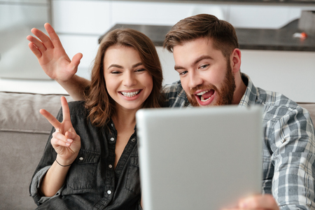 Image of young cheerful loving couple in kitchen at home indoors talking with friends by tablet computer.の写真素材