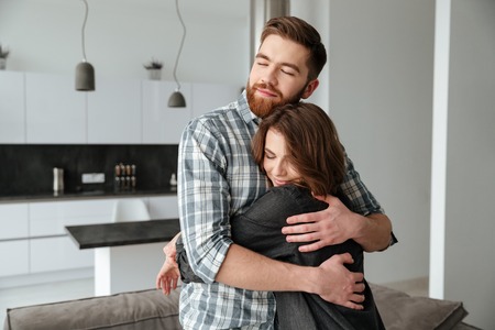 Image of young loving couple standing in kitchen at home indoors. Looking aside while hugging.の写真素材