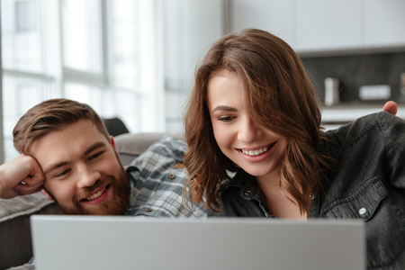 Photo of young cheerful loving couple lies on sofa in kitchen at home indoors using laptop computer.の写真素材