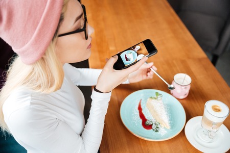 Image of attractive young lady wearing hat and glasses make photo of cake and coffee by phone while sitting in cafe.の写真素材