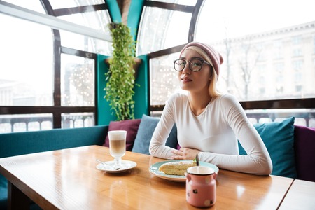 Beautiful young woman in hat and glasses drinking coffee and eating cake in cafeの写真素材
