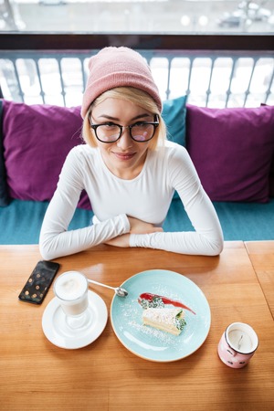 Image of cheerful young woman wearing hat and glasses eating cake and drinking coffee while sitting in cafe.の写真素材