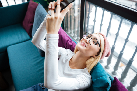 Photo of happy young lady wearing hat and glasses using cellphone while sitting in cafe.の写真素材