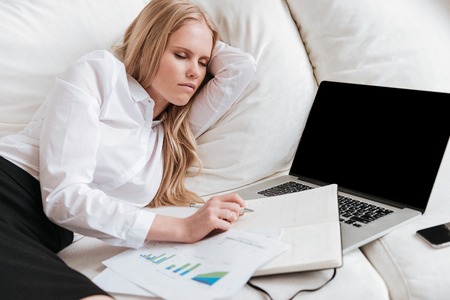 Image of pretty business lady lies in office on sofa. Looking at documents while using laptop.の写真素材