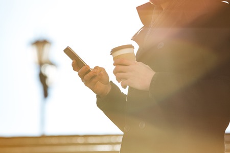 Closeup of man using cell phone and drinking coffee outdoors in sunny dayの写真素材