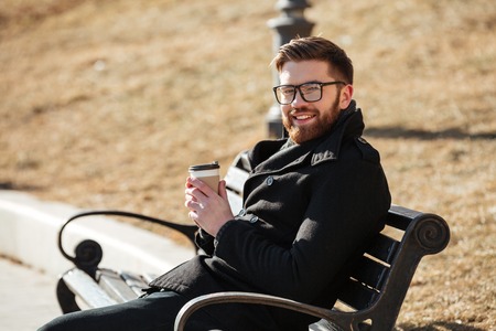 Cheerful bearded young man in glasses sitting on bench and drinking coffeeの写真素材