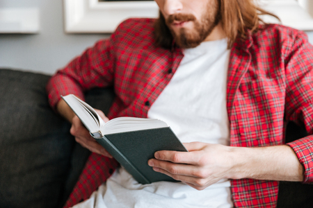 Closeup of bearded young man in plaid shirt reading book at homeの写真素材