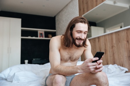 Happy bearded young man using cell phone sitting on bed in bedroomの写真素材