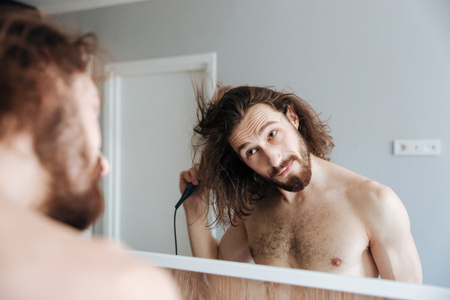 Attractive young man drying hair with hairdryer at homeの写真素材