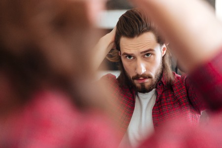 Portrait of attractive bearded young man in plaid shirt standing and looking at the mirrorの写真素材