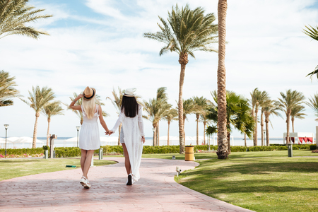 Back view of two carefree young women walking and holding hands on summer resortの写真素材