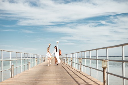 Two happy young women holding hands, walking on pier and looking backの写真素材