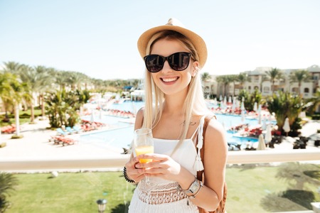 Cheerful young woman in hat and sunglasses drinking juice on summer resortの写真素材