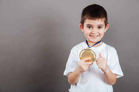 Happy little boy with gold trophy medal showing thumbs up over grey backgroundの写真素材