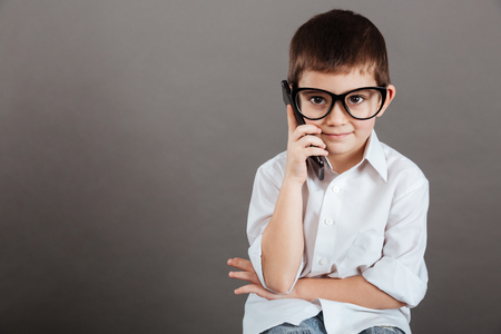 Serious little boy in glasses sitting and talking on mobile phone over grey backgroundの写真素材