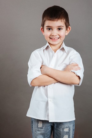 Smiling little boy in white shirt standing with arms crossed over grey backgroundの写真素材