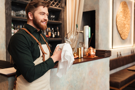 Image of amazing young bearded man bartender standing in cafe. Looking aside and wipe the glass.の写真素材