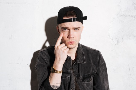 Serious hipster in snap back pointing at his eye and looking at cameraの写真素材